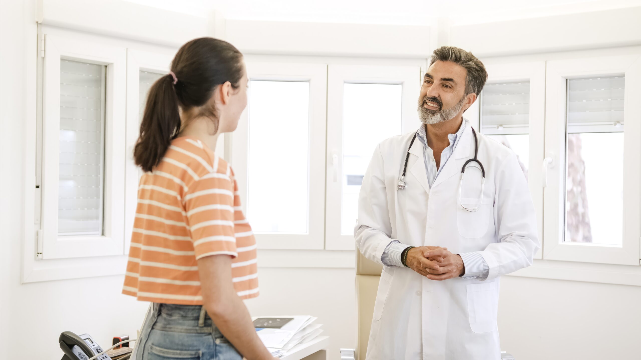 Happy mature male doctor talking to young female patient while standing during consultation in clinic